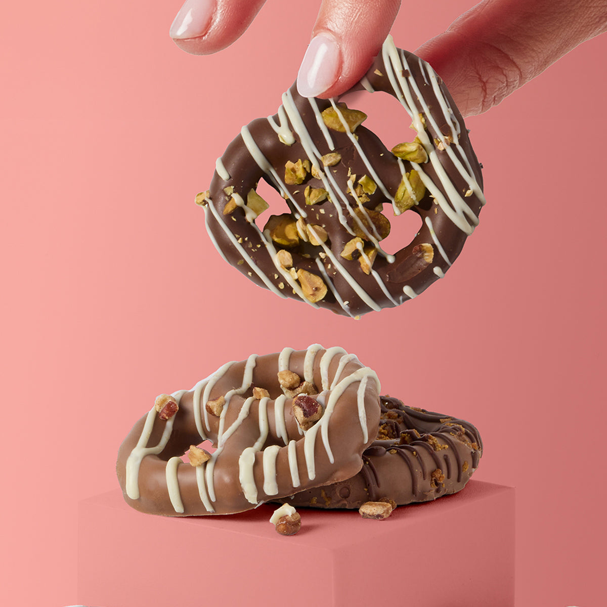 A hand holds a chocolate-covered pretzel above two stacked pretzels on a pink pedestal against a solid pink background. The pretzels are drizzled with white chocolate and topped with chopped pistachios.